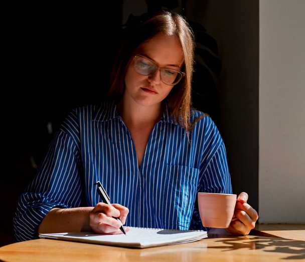 Young smart woman writing plans in diary, sitting at wooden desk in coffee shop with cup and sun light from window - Fotoğraf, Görsel