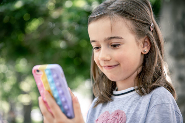 A little girl holds in her hand a phone in a case with pimples pop it, a trendy anti stress toy. - Photo, image