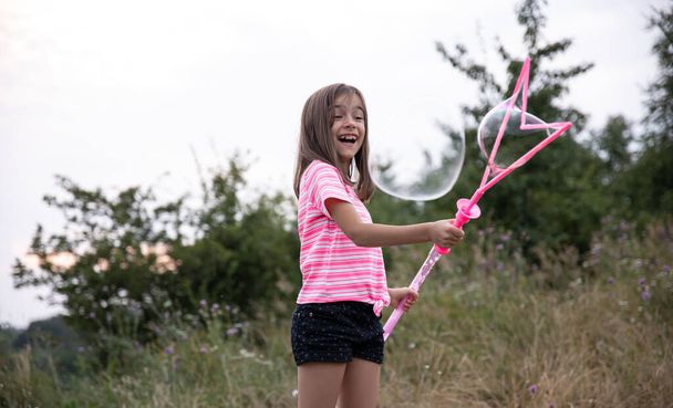 A little funny girl blows soap bubbles in the summer in a field, outdoor summer activities. - Foto, afbeelding