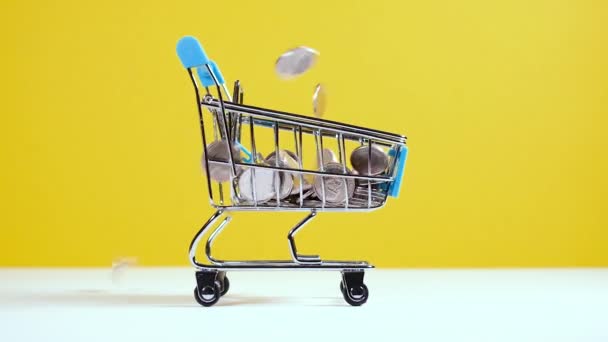Monety wchodzą do koszyka supermarketu zabawki lub wózek izolowane na jasnożółtym tle. Bezpieczne zakupy online lub koncepcja sprzedaży. - Materiał filmowy, wideo