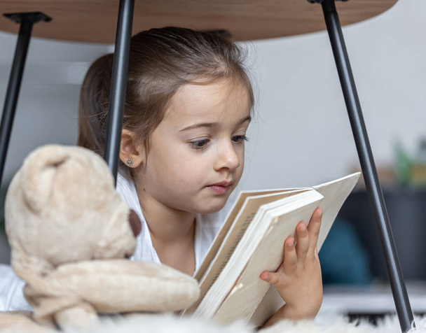 Portrait d'une mignonne petite fille lisant un livre à la maison, allongée sur le sol avec son jouet préféré. - Photo, image