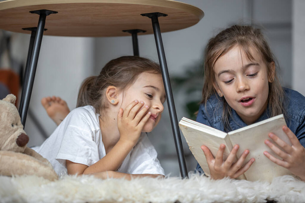 Deux petites sœurs s'amusent à lire un livre ensemble alors qu'elles sont allongées par terre dans leur chambre. - Photo, image