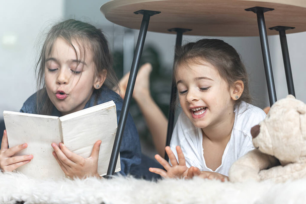 Δύο μικρές αδερφές διαβάζουν ένα βιβλίο ξαπλωμένο στο πάτωμα πάνω σε μια ζεστή κουβέρτα.. - Φωτογραφία, εικόνα