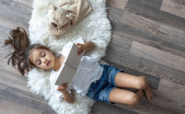 Çocuk evde rahat bir halının üzerinde en sevdiği oyuncak ayısıyla uzanan bir kitap okuyor.. - Fotoğraf, Görsel