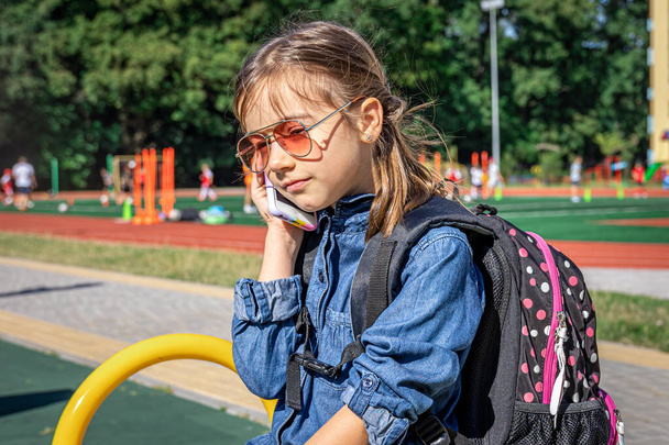 Маленькая девочка, ученица начальной школы в солнцезащитных очках звонит, просто разговаривает, общается. - Фото, изображение
