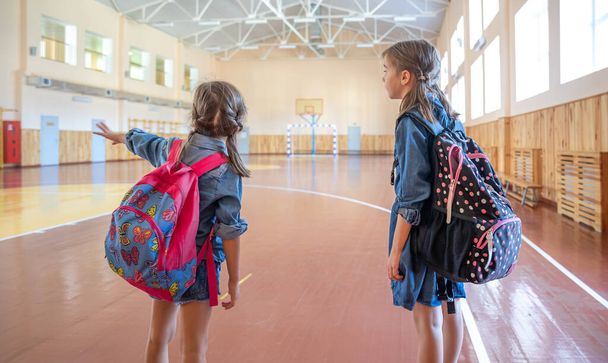 Małe słodkie dziewczynki, uczennice, z plecakami po szkole w pustej sali gimnastycznej szkoły. - Zdjęcie, obraz