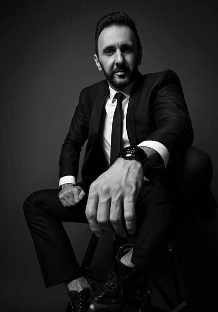 Black and white. Friendly smiling man in modern office suit sits holding hand on his knee, looking down at camera - Foto, Imagem