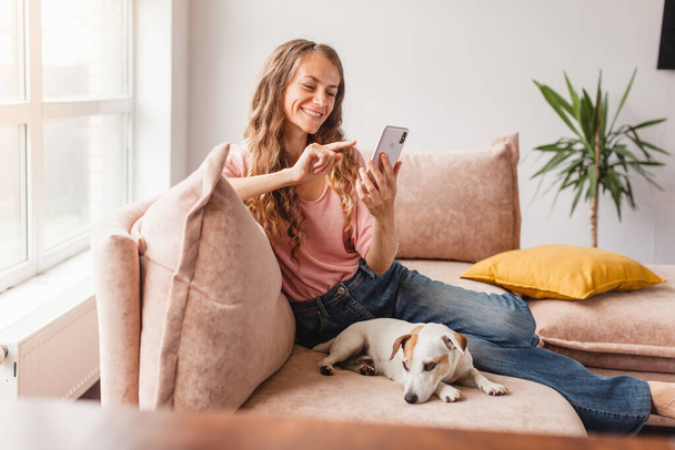 Happy casual beautiful woman is talking on a mobile phone sitting on a sofa at home. Attractive smiling female using smart phone - Photo, image