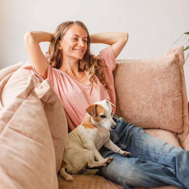 Happy millennial girl with dog sit relax on comfortable couch in living room hands over head, peaceful young woman rest on cozy sofa at home take nap dream with closed breathe fresh air, stress free concept - Fotó, kép