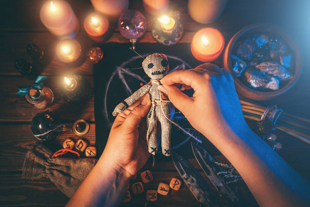 Tovenares of heks prikt naalden in voodoo pop aan rituele tafel met pentagram, brandende kaarsen en andere occulte voorwerpen, bovenaanzicht. Voodoo hekserij, spiritualiteit en occultisme concept - Foto, afbeelding