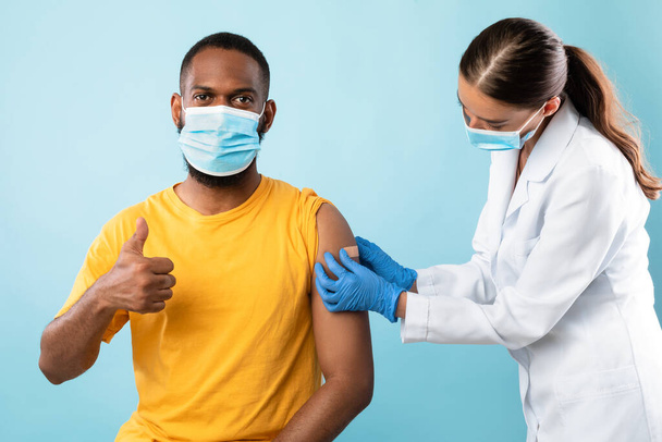 Black guy in face mask showing thumb up gesture after covid vaccine shot, recommending anti coronavirus vaccination - Foto, imagen