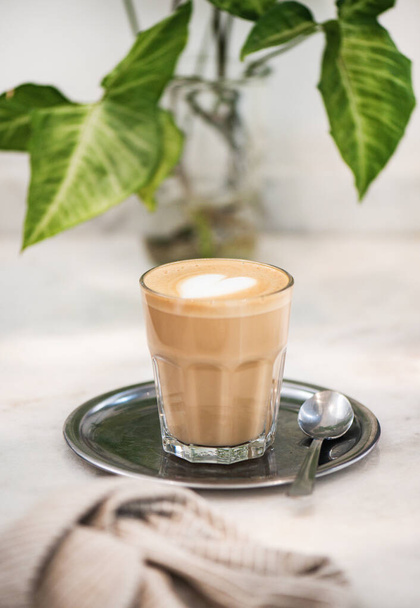 Glass of flat white coffee with heart shaped foam milk and spoon on silver tray and green plant on background, selective focus. Idyllic drink for breakfast concept - Valokuva, kuva
