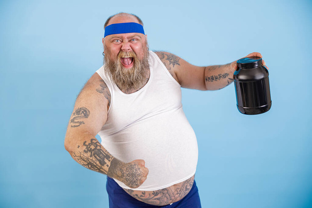 Hombre barbudo alegre con sobrepeso sostiene botella negra de suplemento de proteína sobre fondo azul claro - Foto, imagen