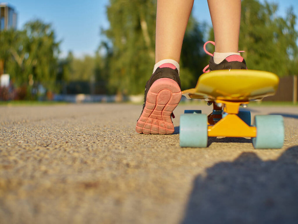 Dziewczyna dzieciak stojący na asfaltowej drodze i żółty penny pokładzie z niebieskimi kołami na tle ulicy w czasie zachodu słońca. - Zdjęcie, obraz