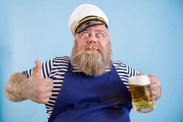 Radosny grubas w marynarskim garniturze trzyma piwo i pokazuje kciuk w górę na jasnoniebieskim tle - Zdjęcie, obraz