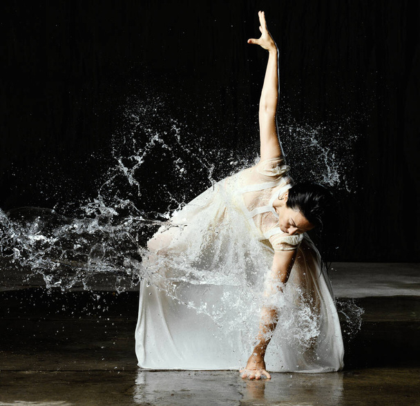 schöne Frau kaukasischen Aussehens mit schwarzem Haar tanzt in Wassertropfen auf schwarzem Hintergrund. Die Frau trägt ein weißes Chiffonkleid - Foto, Bild