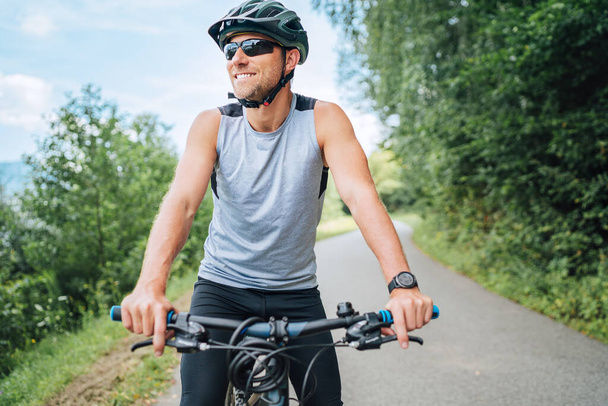 Half-length portrait of a sincerely smiling man dressed in cycling clothes, helmet and sunglasses riding a bicycle on the asphalt out-of-town bicycle path. Active sporty people concept image. - 写真・画像