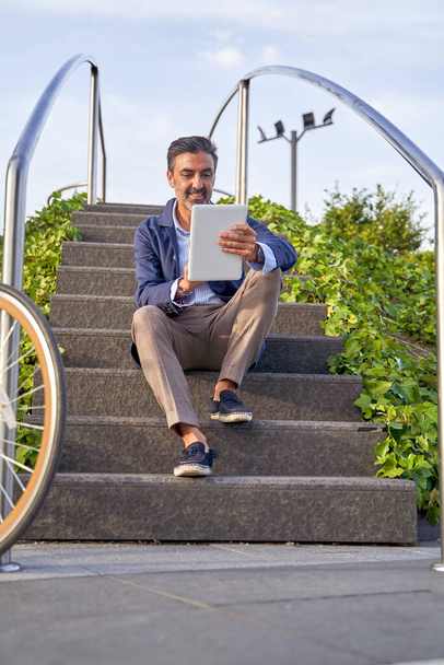 Empresario sentado en una escalera frente a un edificio de oficinas, leyendo un correo electrónico en una tableta digital. El hombre monta en bicicleta por la ciudad.  - Foto, imagen