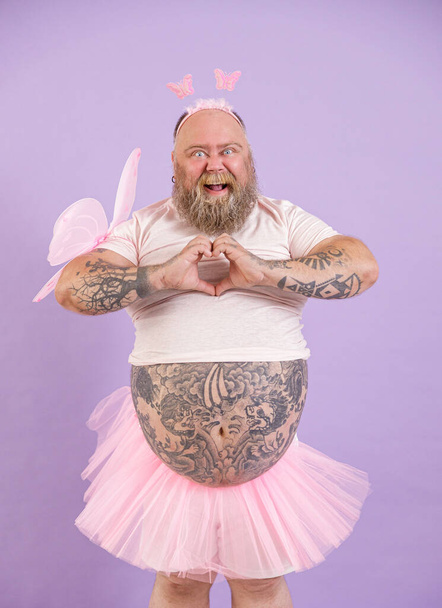 Joyful plus size man in fairy suit shows heart with hands on purple background - Фото, изображение