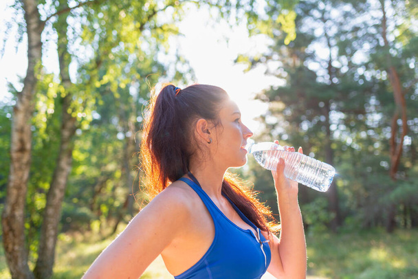 Dívka pije vodu z láhve v lese. Mladá žena držící láhev vody v lese, portrét. pozitivní dáma ve sportovním oblečení si odpočinout po běhu, běhání. v slunečný letní den - Fotografie, Obrázek