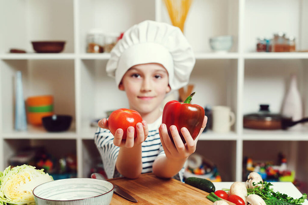Küçük şef yemek yapmak için peper ve domates tutuyor. Şef şapkası ve üniforması olan sevimli bir çocuk. Yemek pişirme konsepti. Oğlum aile yemeği için sağlıklı yemek hazırlıyor. Çocuk profesyonel şef olmak istiyor.. - Fotoğraf, Görsel