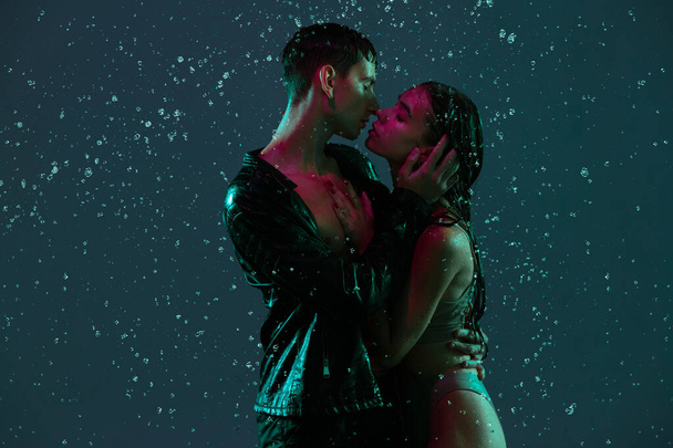 Junges Paar, Mann und Frau umarmen sich, küssen sich im Regen vor dunklem Hintergrund im Neonlicht. - Foto, Bild