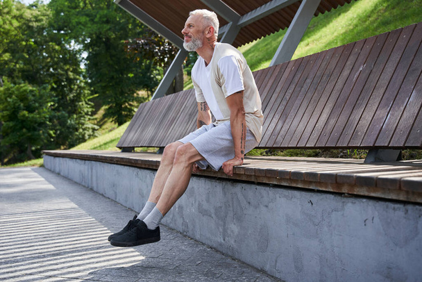 Senior man sitting at the wooden bench and looking away while resting - Zdjęcie, obraz