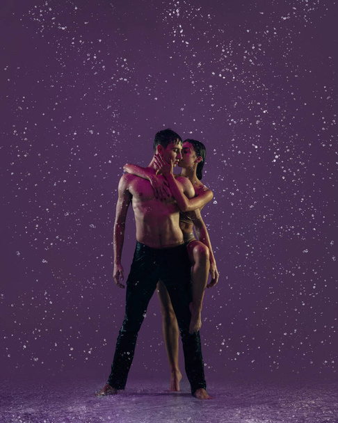 Two young ballet dancers, man and woman dancing in the raindrops over purple background. Love, art, inspiration concept. - Photo, Image