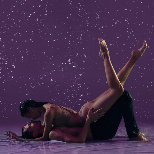 Two young ballet dancers, man and woman lying on floor under the rain over purple background. Love, art, inspiration concept. - Photo, Image