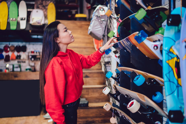 Side view of attractive teenage woman choosing longboard from store rack during Black Friday sales, beautiful Japanese hipster girl spending day for buying skateboard and practicing hobby skills - Foto, immagini