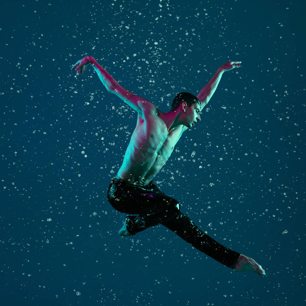 One young male ballet dancer in motion, action under the rain over blue background in neon light. - Foto, Imagem