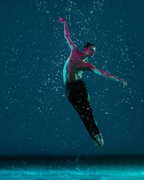 One young male ballet dancer in motion, action under the rain over blue background in neon light. - Foto, Imagen