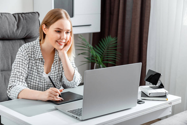 Smiling happy Woman designer using graphic tablet and laptop for work in home office. Freelance retoucher illustrator young caucasian blonde woman working remotely from home workspace. - Foto, Imagem