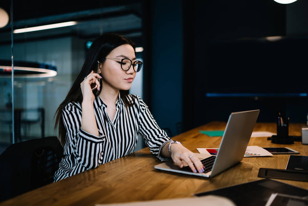 Dark haired young focused female with glasses and casual shirt talking on phone and using laptop while sitting at table in modern office - Photo, Image