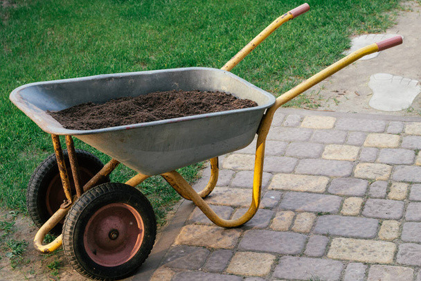 Wózek z glebą na kostki brukowej obok zielonego trawnika. Taczka z ziemią do ogrodnictwa - Zdjęcie, obraz