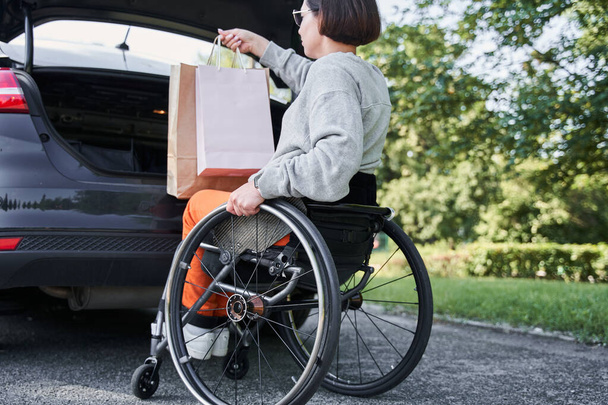 Girl sitting at the wheelchair is putting bags with purchases into car trunk - Foto, afbeelding