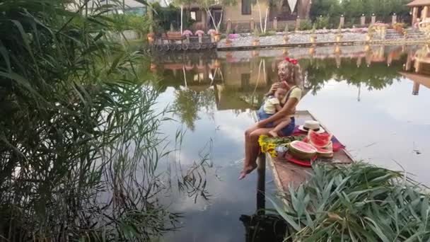 Mom and little daughter, dressed in denim shorts with suspenders, with red butterflies on their heads, sit on a fishing platform on the river and hug  - Footage, Video