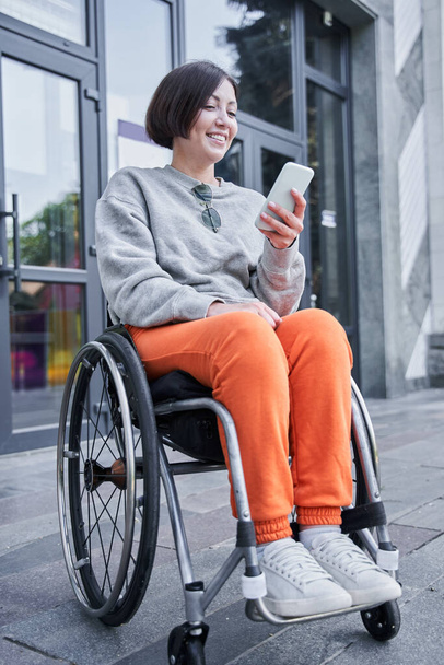Girl with lower body disability sitting at the wheelchair and looking at her smartphone - Foto, immagini