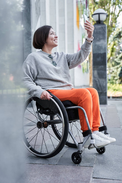 Дівчина з втратою функції ноги робить селфі на смартфоні, сидячи на інвалідному візку
 - Фото, зображення