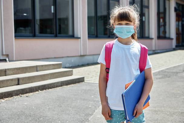De vuelta a la escuela. Retrato de una chica con una mochila en una máscara médica en el patio de la escuela. Proteger a los niños de virus e infecciones durante una epidemia de gripe - Foto, imagen