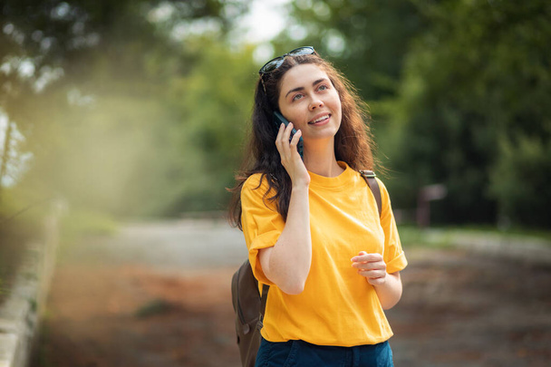 Портрет молодой улыбающейся женщины, разговаривающей по телефону. Улица или парк на заднем плане. - Фото, изображение