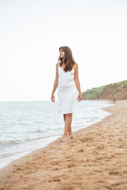girl in a white dress on the beach near the sea, beautiful sunset, smiling - Фото, изображение