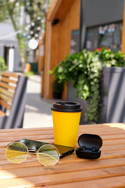 Ein Glas Kaffee mit modernen drahtlosen Kopfhörern und ein Smartphone liegen auf einem Tisch in einem Café im Freien. Arbeit im Freien. Freizeit für Erholung - Foto, Bild