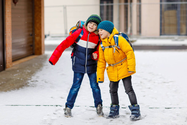 Deux petits garçons de classe primaire marchant jusqu'à l'école pendant les chutes de neige. Enfants heureux s'amuser et jouer avec la première neige. Frères et sœurs et meilleurs amis avec sac à dos en vêtements d'hiver colorés. - Photo, image