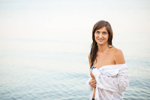 meisje in een badpak en wit shirt op het strand in de buurt van de zee, prachtige zonsondergang, glimlachen - Foto, afbeelding
