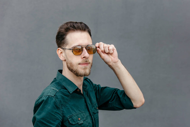 Portrait of a young man wearing sunglasses on a gray background touching his glasses - Photo, Image