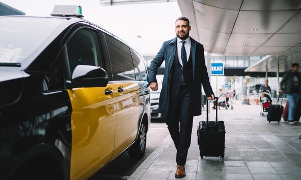Confident successful businessman in formal wear holding baggage and looking forward while walking to taxi on street near airport terminal - Photo, Image