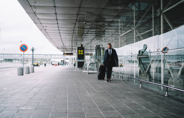 Full length of young man in formal suit with luggage walking along road against large airport building with glass walls - Photo, image