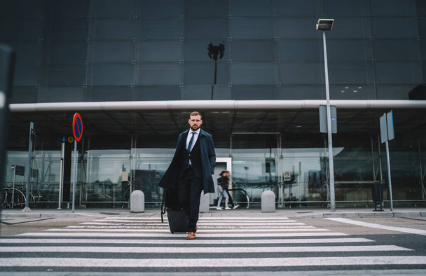 Confident manager wearing formal clothes with luggage looking at camera while walking on crosswalk to transport in  contemporary city - Photo, image