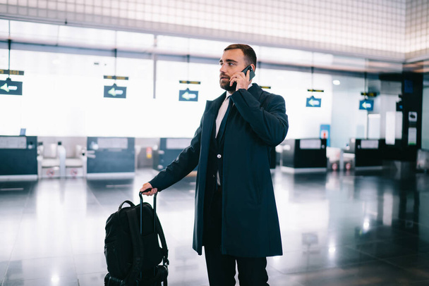 Side view of elegant serious male in black suit with suitcase standing in airport hall while talking on mobile phone - Zdjęcie, obraz